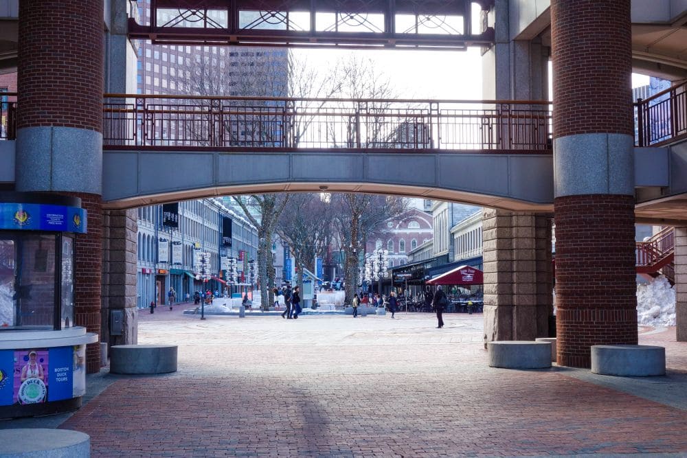 Ein gemauerter Torbogen rahmt den Blick auf einen Bostoner Stadtplatz ein, wo die Menschen unter freiem Himmel an Sitzgelegenheiten und historischen Gebäuden vorbeischlendern.