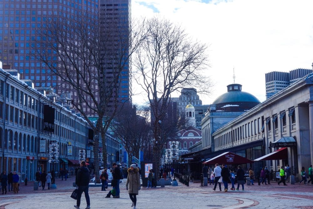 Ein Spaziergang über einen kopfsteingepflasterten Weg mit historischen Gebäuden und Geschäften im Bostoner Stadtteil Quincy Market, mit blattlosen Bäumen und modernen Wolkenkratzern vor einem wolkenverhangenen Bostoner Himmel.