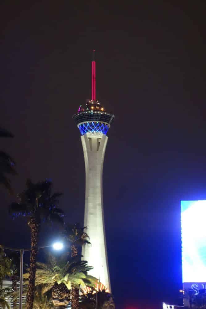 Der Stratosphere Tower in Las Vegas ist nachts mit roten, blauen und weißen Lichtern, Palmen im Vordergrund und einer leuchtenden Werbetafel auf der rechten Seite beleuchtet.