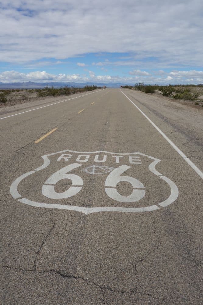 Eine lange, gerade Strecke der Route 66 in Illinois mit dem auf die Straße gemalten Emblem, umgeben von Wüstenlandschaft und niedrigen Büschen unter einem teilweise bewölkten Himmel.