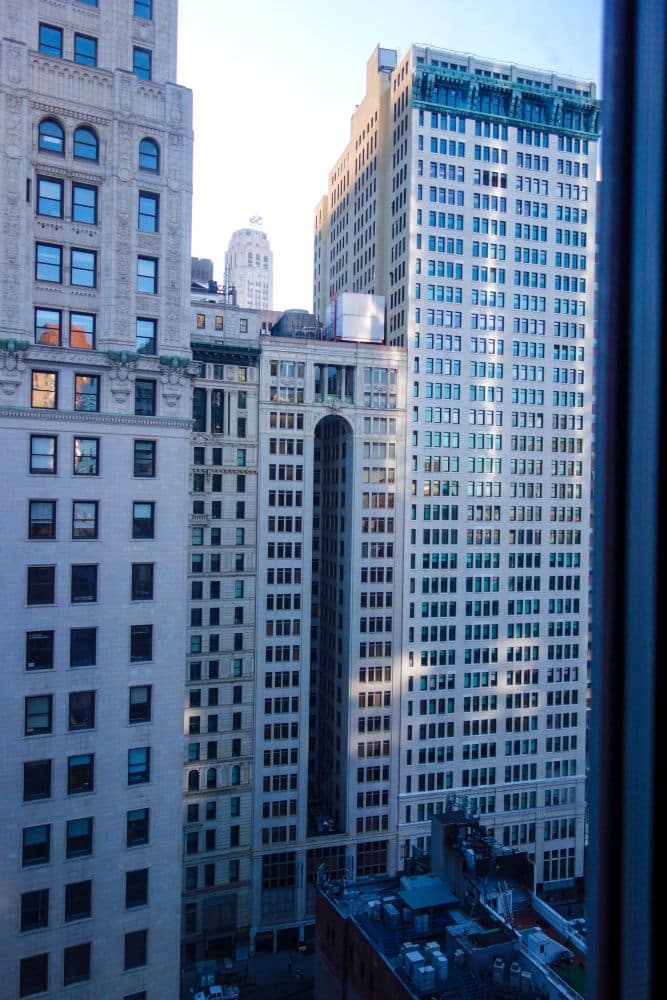 Ein Blick auf mehrere hohe Bürogebäude mit vielen Fenstern in einer Stadt, von einem hohen Aussichtspunkt aus gesehen; darüber ist der blaue Himmel zu sehen, und das Sonnenlicht spiegelt sich in den Glas- und Steinfassaden.