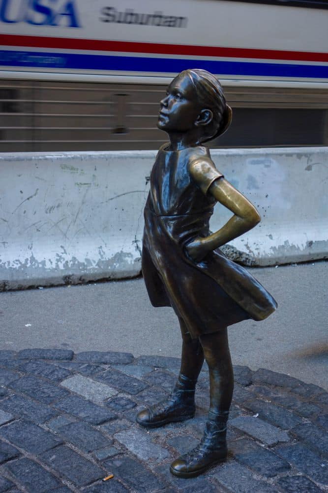 Die Bronzestatue eines jungen Mädchens steht selbstbewusst, die Hände in die Hüften gestemmt, in einem Finanzviertel und blickt nach oben. Sie trägt ein Kleid und Stiefel. Ein unscharfes Fahrzeug und eine Betonbarriere befinden sich im Hintergrund auf einer Stadtstraße im Financial District.