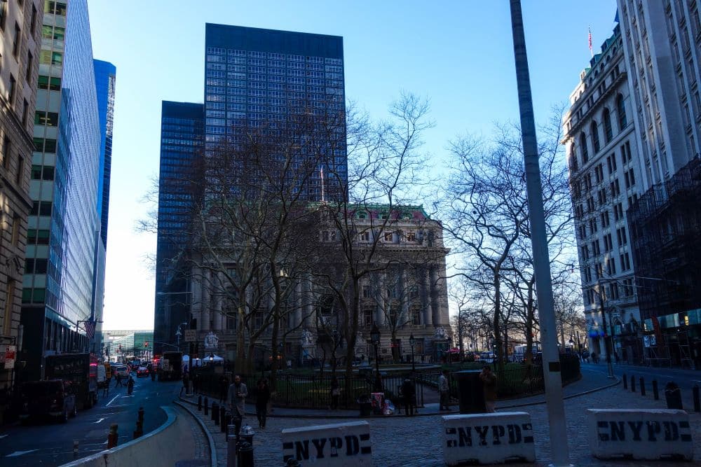 Eine Straßenszene in New Yorks Financial District mit hohen Gebäuden, blattlosen Bäumen und NYPD-Sperren im Vordergrund. Der Himmel ist klar und blau, und im Hintergrund sind Fußgänger und Autos zu sehen.