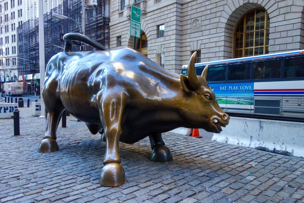 Bronzestatue des "Charging Bull" auf der Kopfsteinpflasterstraße im New Yorker Financial District als Symbol für finanzielle Stärke, mit hohen Gebäuden und einem Bus im Hintergrund.