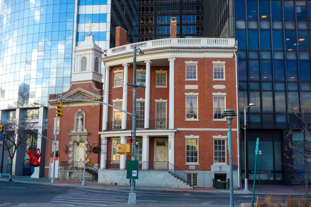 Ein historisches Gebäude aus rotem Backstein mit weißen Säulen und einer angebauten Kirche steht an einer Straßenecke im Herzen des Financial District, umgeben von modernen gläsernen Wolkenkratzern, Ampeln und Straßenschildern.