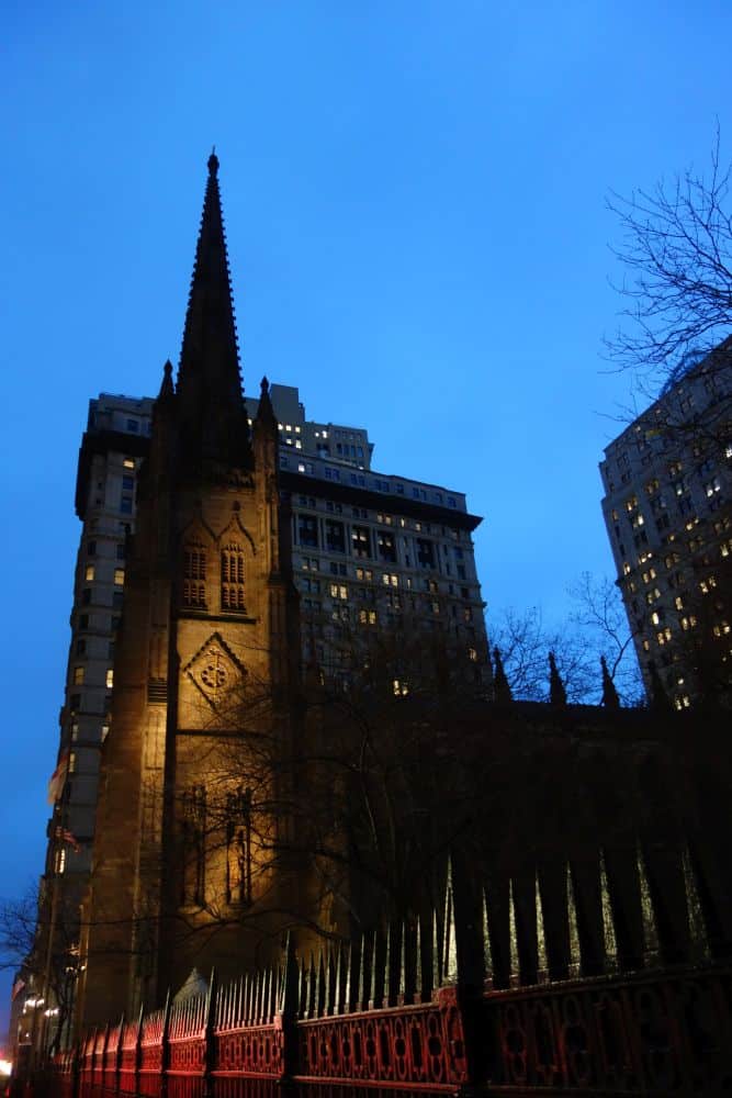 Eine gotische Kirche mit einem hohen Kirchturm ist in der Abenddämmerung beleuchtet, umgeben von kahlen Bäumen und modernen Gebäuden im Finanzviertel der Stadt. Im Vordergrund ist ein dekorativer Eisenzaun mit spitzen Zacken zu sehen.