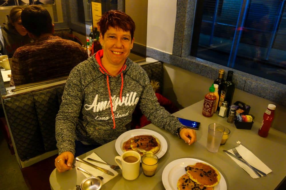 Eine Frau in einem grauen Amityville Long Island-Kapuzenpulli sitzt an einem Imbissstand und lächelt in die Kamera. Sie genießt Pfannkuchen, Kaffee und Orangensaft - ein einfaches Vergnügen, das beweist, dass ein toller Morgen nicht mit einem finanziellen Kraftakt verbunden sein muss.