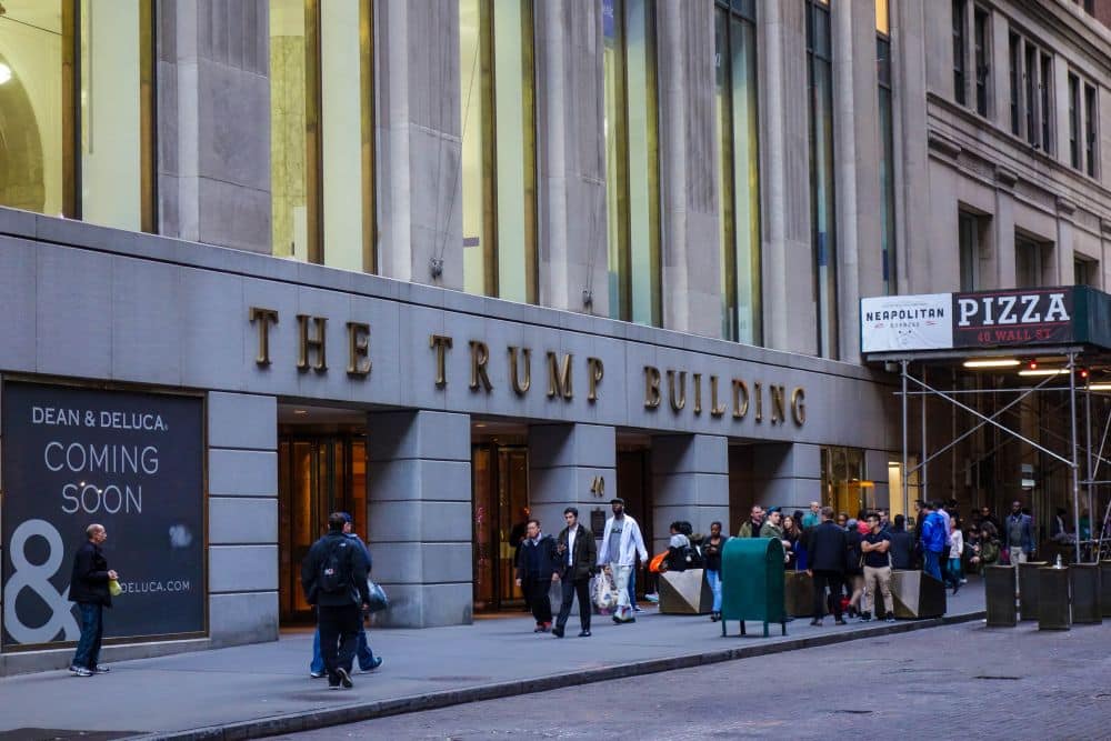 Menschen stehen und gehen vor dem Trump Building, einem hohen Finanzgebäude mit großen Fenstern an einer belebten Straße in der Stadt. Ein Schild für ein neues Geschäft und eine Pizzeria sind in der Nähe zu sehen.