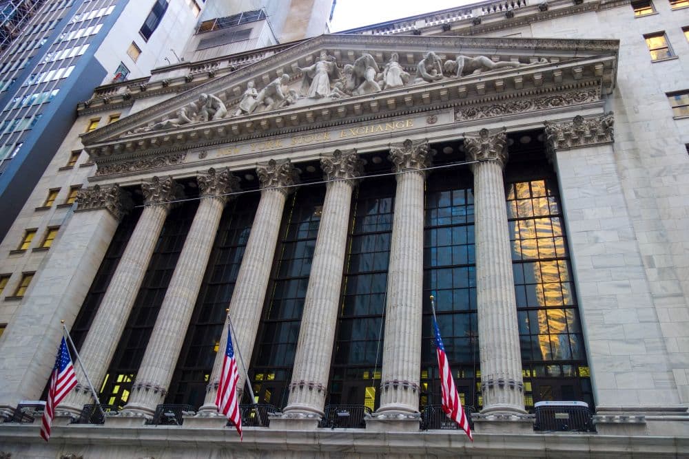 Das Bild zeigt die Finanzfassade der New Yorker Börse mit hohen Säulen, einem dreieckigen Giebel mit Skulpturen und drei amerikanischen Flaggen im Vordergrund. Das Gebäude ist von modernen Hochhäusern umgeben.