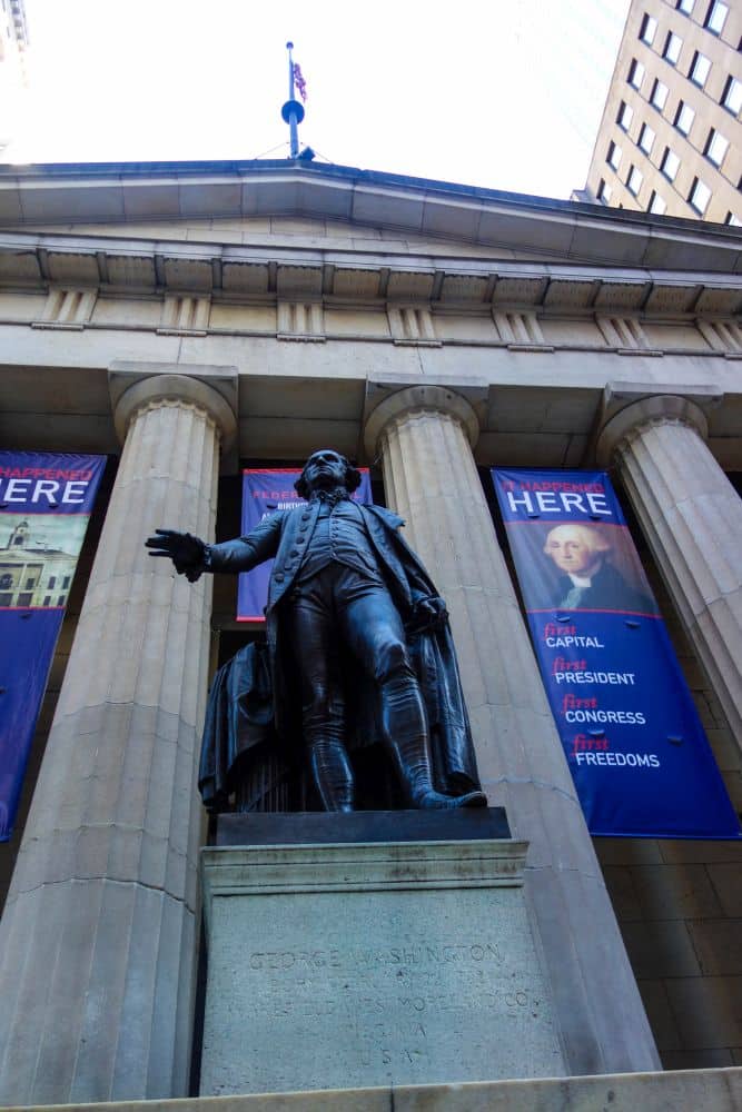Eine Bronzestatue von George Washington steht vor einem Säulengebäude mit Bannern zum Thema Finanzen, auf denen "HIER" steht und sein Porträt neben Wörtern wie "CAPITAL", "PRESIDENT", "CONGRESS" und "FREEDOMS" zu sehen ist.
