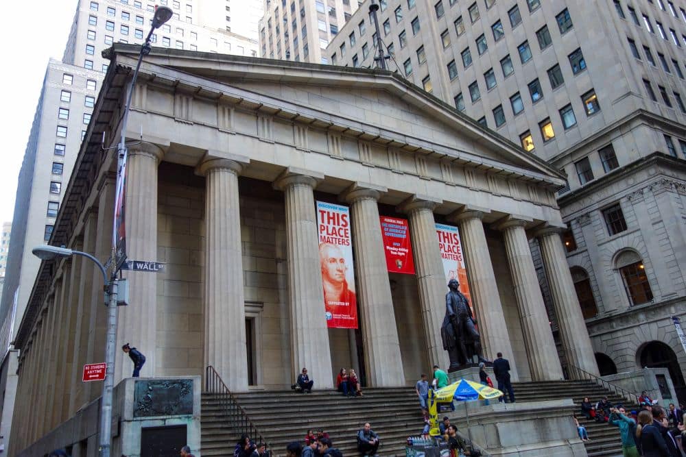 Ein historisches Finanzgebäude mit hohen Säulen und Bannern mit der Aufschrift This Place Matters hängt am Eingang. Menschen sitzen auf den Stufen, und eine Statue steht davor. Die Szene befindet sich in einer Stadt, die von hohen Bürogebäuden umgeben ist.