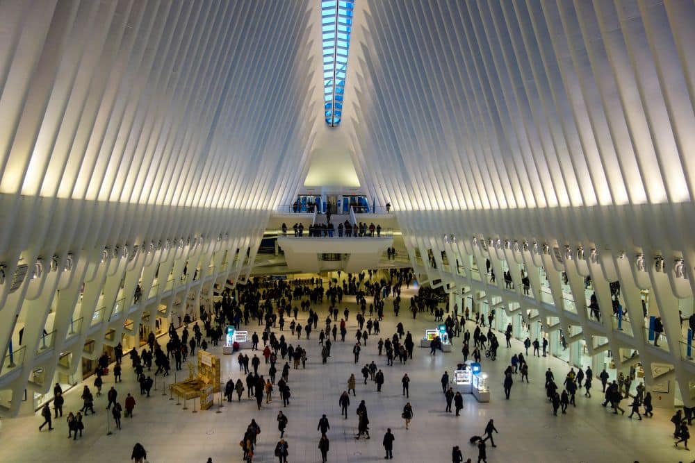 Eine große, helle Halle mit einer hohen Gewölbedecke und einer gerippten Architektur, die an ein modernes Finanzzentrum erinnert; die Menschen gehen umher und versammeln sich in weißen Räumen und auf mehreren Ebenen.