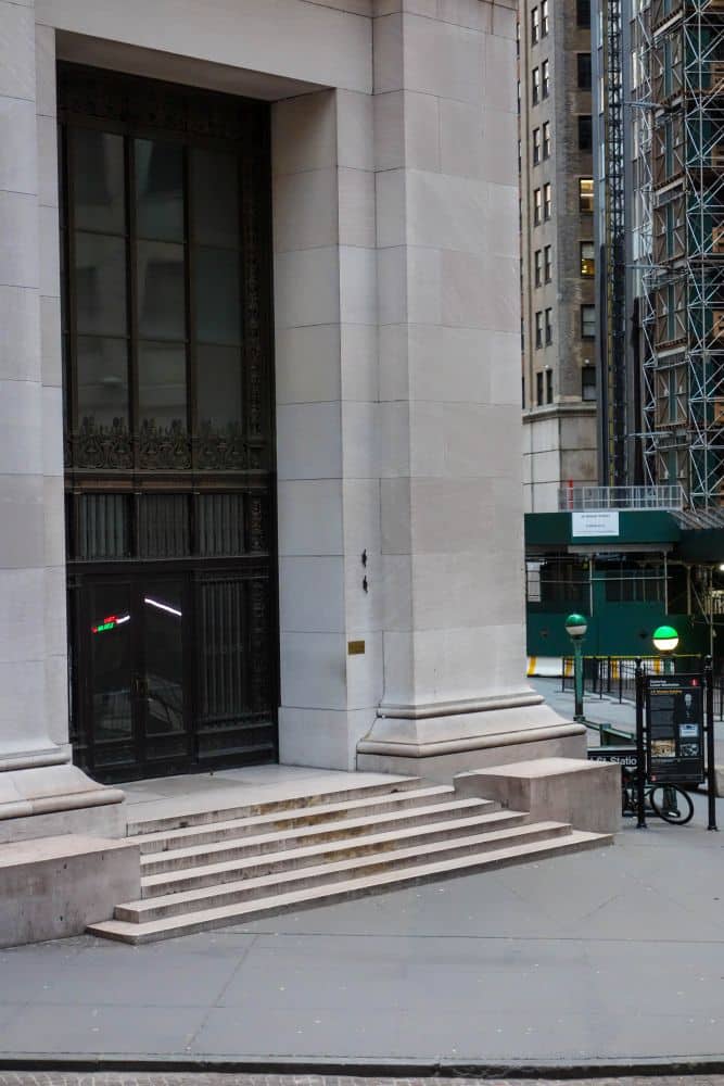 Steinstufen führen zum Eingang eines großen Finanzgebäudes mit hohen Säulen und dunklen Türen. In der Nähe sind grüne U-Bahn-Eingangsleuchten und Baugerüste in einer ruhigen Stadtstraße zu sehen.