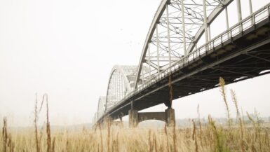 Eine große Stahlbogenbrücke spannt sich über ein grasbewachsenes Feld, mit hohem, trockenem Gras im Vordergrund und einem dunstigen Himmel im Hintergrund. Die Struktur der Brücke in Illinois ist aus einem niedrigen Winkel sichtbar.