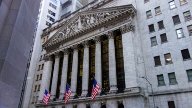 Die vordere Fassade der New Yorker Börse im Financial District mit hohen Säulen, einem dekorativen Giebel und drei amerikanischen Flaggen, umgeben von hohen Gebäuden in Downtown Manhattan.