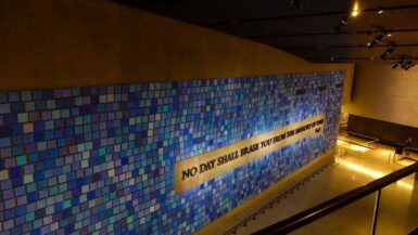 An einer mit blau getönten Kacheln bedeckten Wand steht der Spruch "Kein Tag wird dich aus dem Gedächtnis der Zeit löschen" in einer museumsähnlichen Umgebung mit Scheinwerfern und einem Geländer im 9/11 Museum.