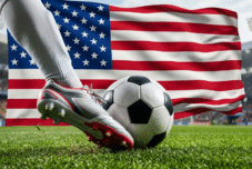 Der Fuß eines Fußballspielers mit grauen und roten Stollen bereitet sich darauf vor, einen schwarz-weißen Fußball auf einem Feld zu kicken, während im Hintergrund des Stadions eine große amerikanische Flagge weht.