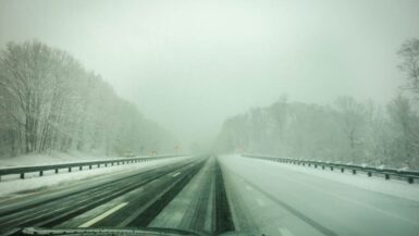 Eine schneebedeckte Autobahn führt durch eine winterliche Landschaft. Die Bäume auf beiden Seiten sind mit Schnee bedeckt und die Sicht ist durch Nebel eingeschränkt. Die Straße scheint vereist und weitgehend leer zu sein.