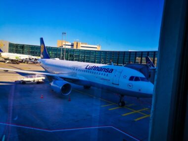 Ein Lufthansa-Flugzeug steht an einem sonnigen Tag an einem Flughafen-Gate. Im Hintergrund sind das Terminalgebäude und eine weitere Lufthansa-Maschine zu sehen.