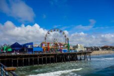 Santa Monica Pier mit einem Riesenrad und bunten Fahrgeschäften über dem Meer, Wellen, die gegen den Pier schlagen, und ein strahlend blauer Himmel mit vereinzelten Wolken im Hintergrund.